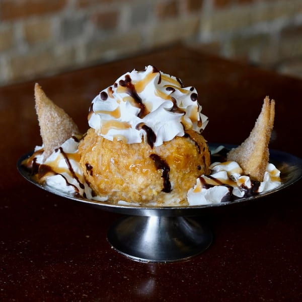 Fried ice cream dessert with whipped cream, chocolate, and caramel drizzles, served with two churro pieces.
