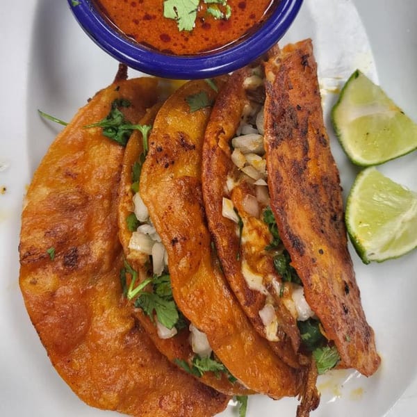 Three El Mariachi birria tacos with cilantro, onions, and lime wedges, served with a side of consommé.