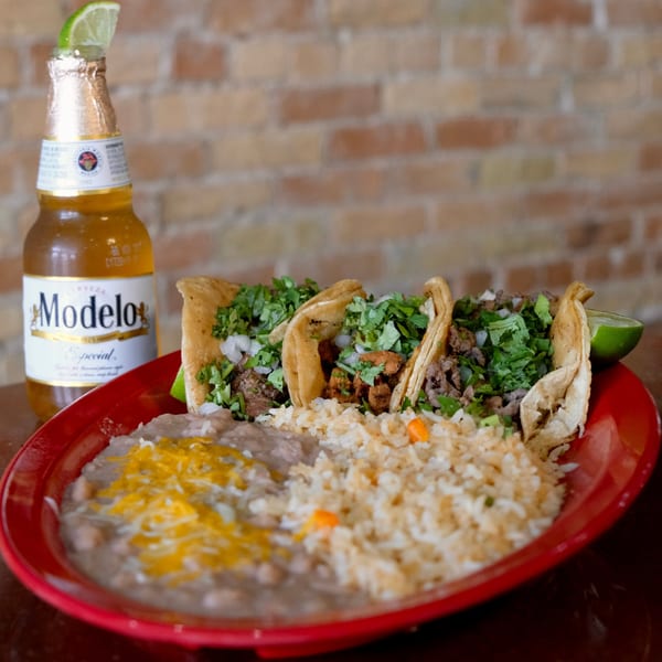 Three tacos with cilantro and onions, rice, and refried beans with cheese, next to a Modelo beer.