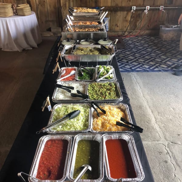A long buffet table laden with Mexican food, including hot dishes, salsas, guacamole, and toppings.